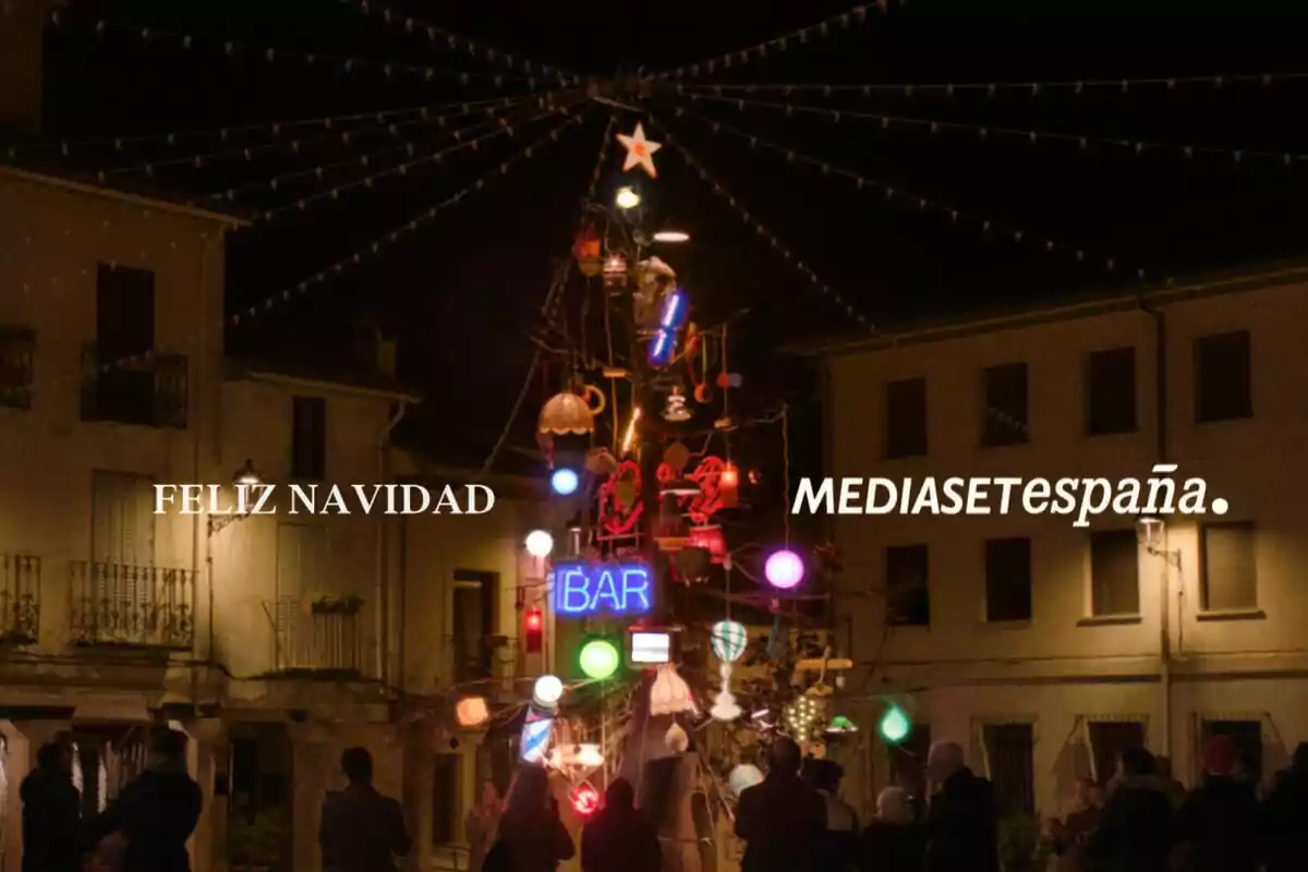 Una plaça il·luminada amb un arbre de Nadal decorat amb llums i adorns colorits, envoltat de persones, amb les paraules "Feliz Navidad" i "Mediaset España" al cel nocturn.