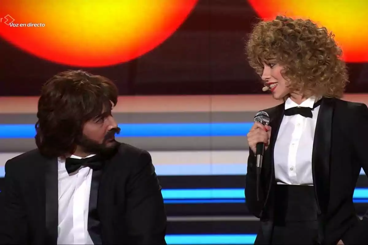 Jorge González i Melani vestides de gala són en un escenari de Tu Cara Me Suena, una d'elles sosté un micròfon mentre l'altra l'observa. Jorge González i Melani vestides de gala són en un escenari de Tu Cara Me Suena, una d'elles sosté un micròfon mentre l'altra l'observa.