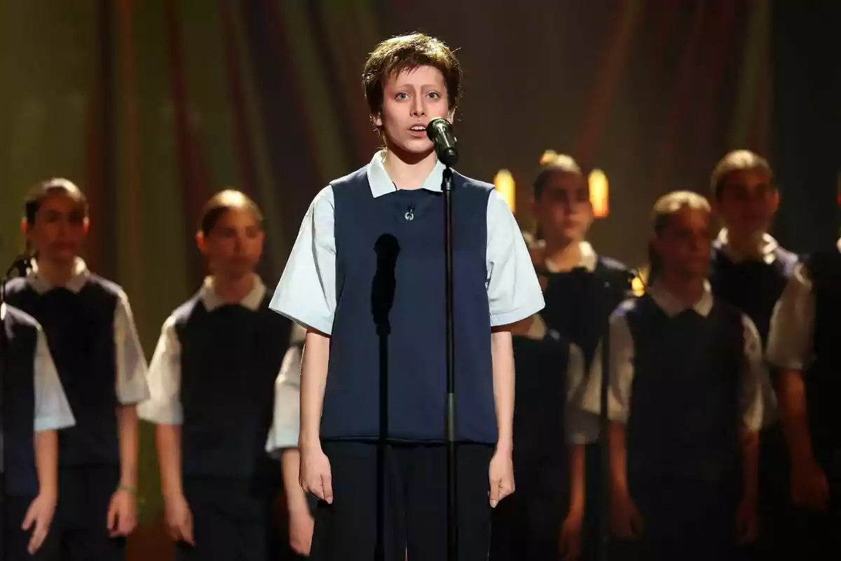Melani vestida amb uniforme escolar canta davant d’un micròfon mentre un grup d’estudiants l’acompanya a l’escenari il·luminat a Tu Cara Me Suena. Melani vestida amb uniforme escolar canta davant d’un micròfon mentre un grup d’estudiants l’acompanya a l’escenari il·luminat a Tu Cara Me Suena.