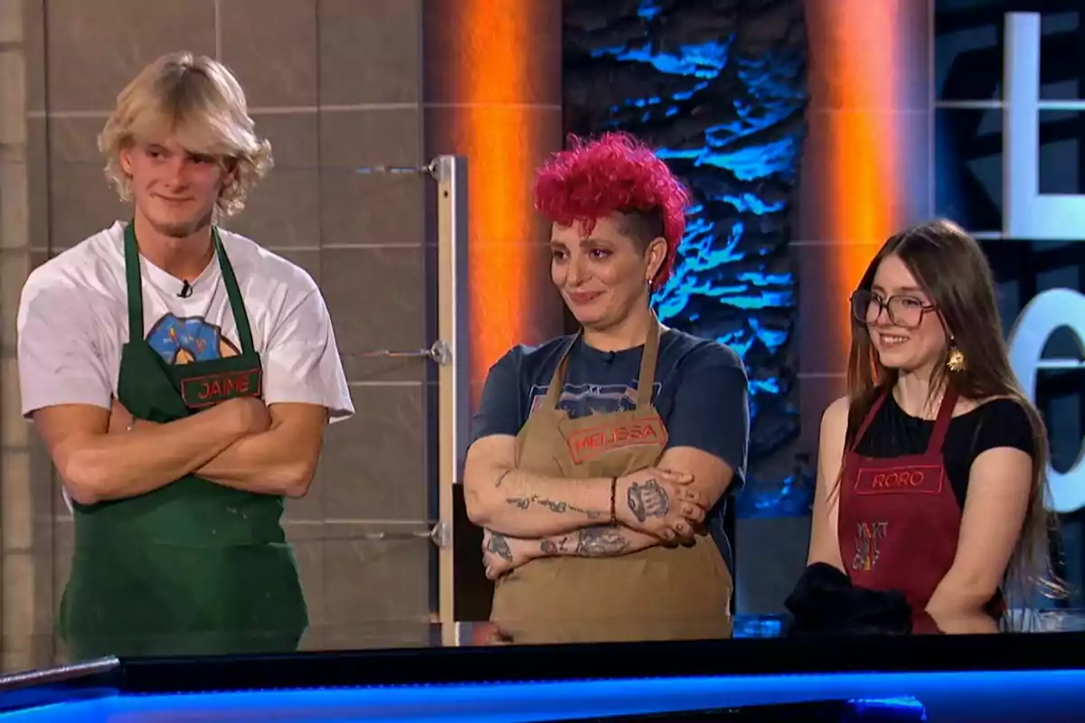 Jaime, Melissa y Roro Bueno con delantales de colores diferentes están de pie, sonriendo y cruzando los brazos en un entorno que parece ser un programa de cocina en Next Level Chef.