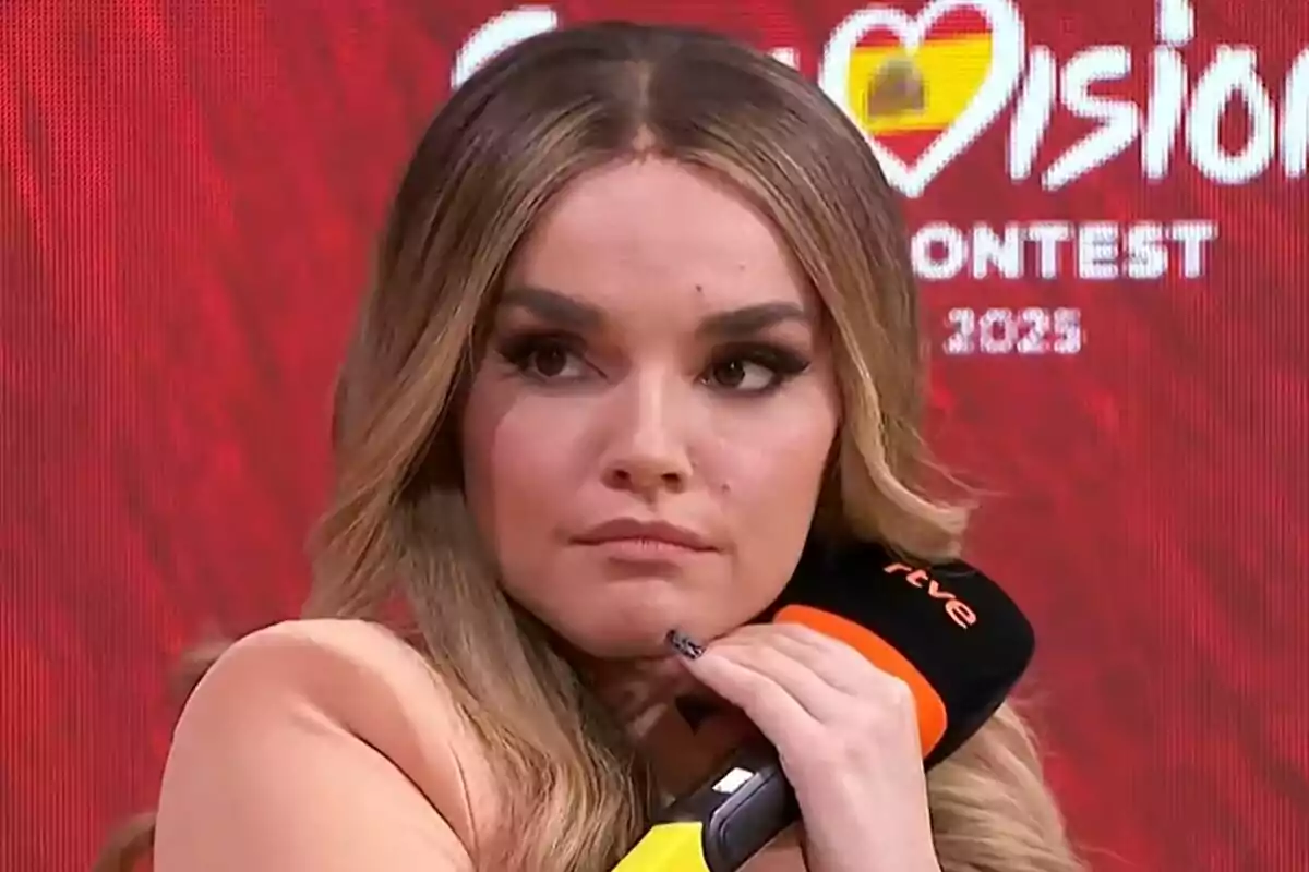 Melody de cabell llarg i ros sostenint un micròfon negre i taronja amb fons vermell i lletres blanques d’un concurs de música d’Eurovisió.