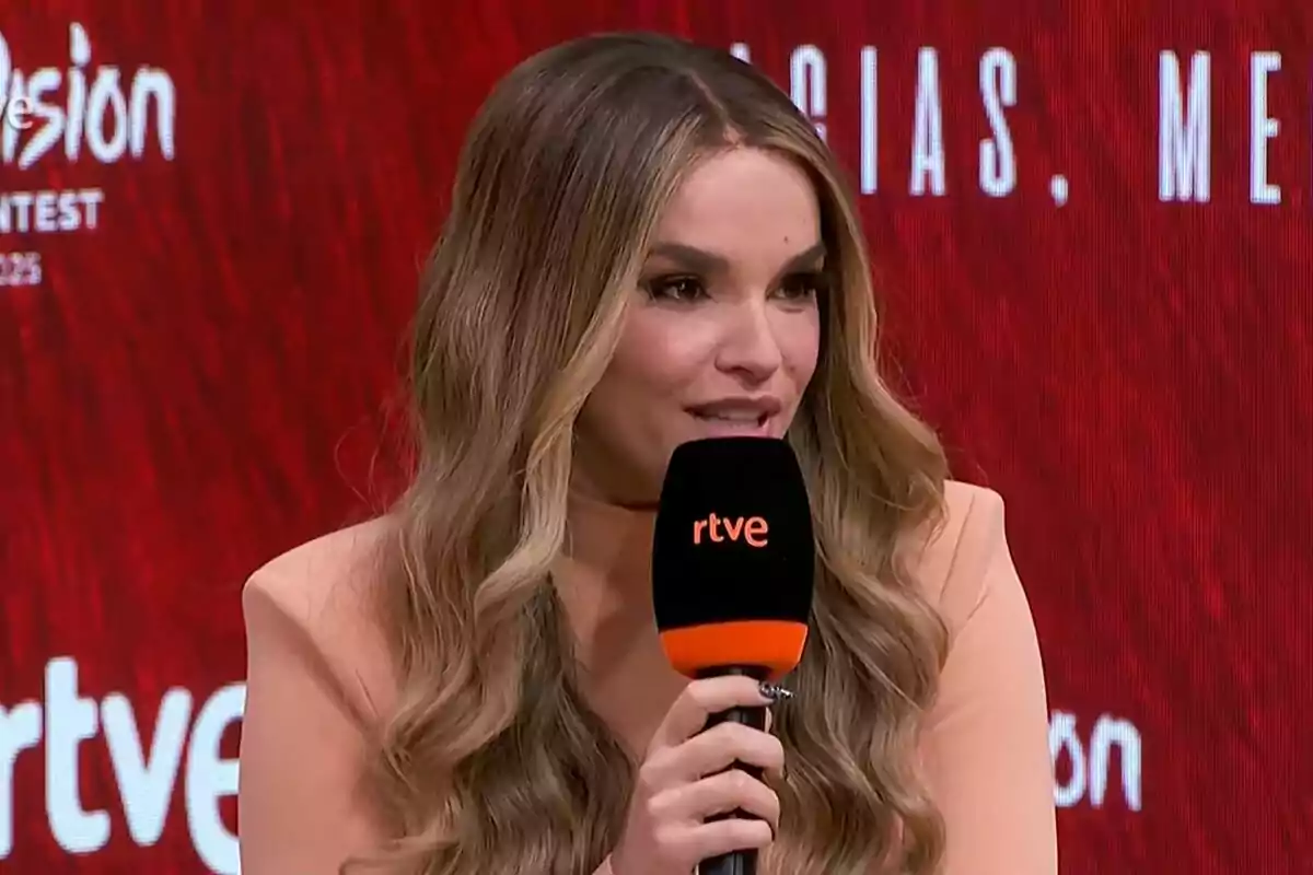 Melody de cabell llarg i ondulat parlant davant d’un micròfon de rtve amb fons vermell i lletres blanques a Eurovisió. Melody de cabell llarg i ondulat parlant davant d’un micròfon de rtve amb fons vermell i lletres blanques a Eurovisió.