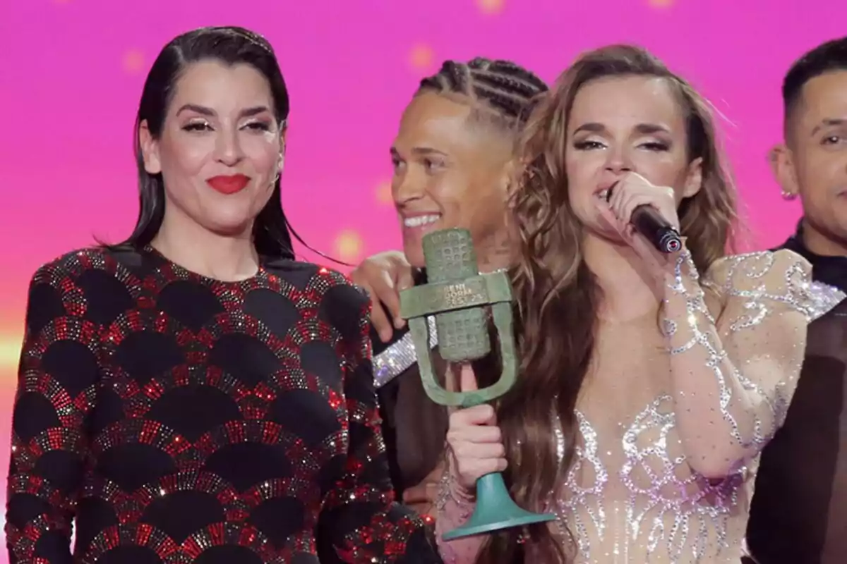 Un grupo de personas en un escenario del Benidorm Fest, Melody sostiene un trofeo y un micrófono junto a Ruth Lorenzo, con un fondo de color rosa brillante.