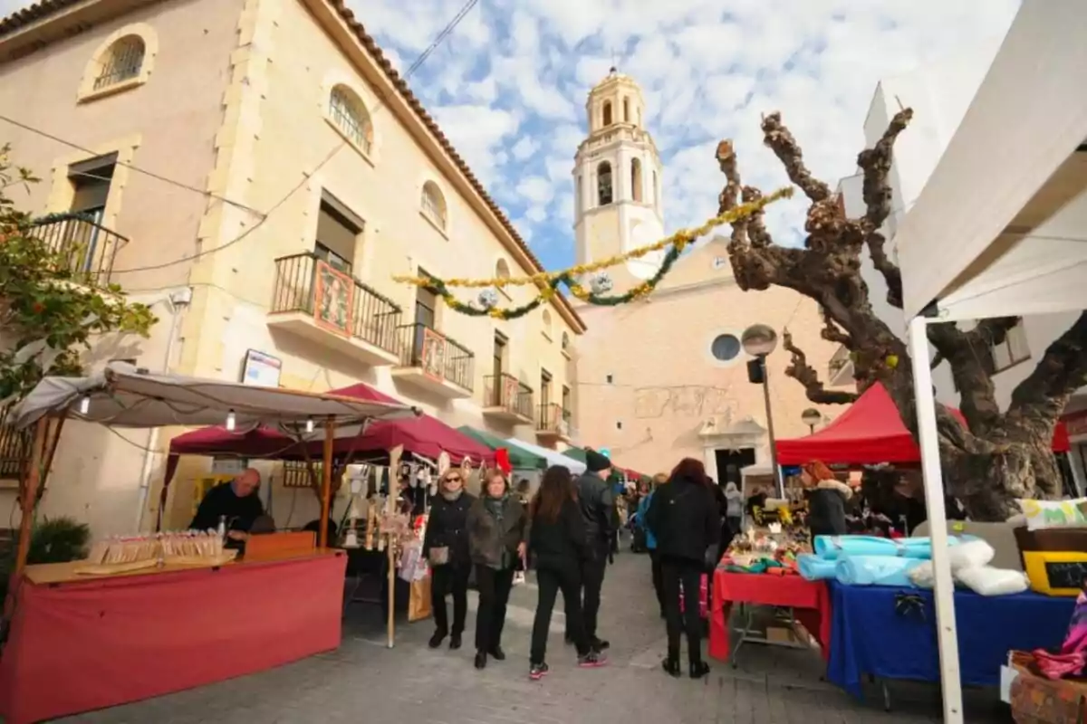 Una fira exterior amb parades de venda i persones caminant en una plaça amb un edifici antic i una torre desglésia al fons.