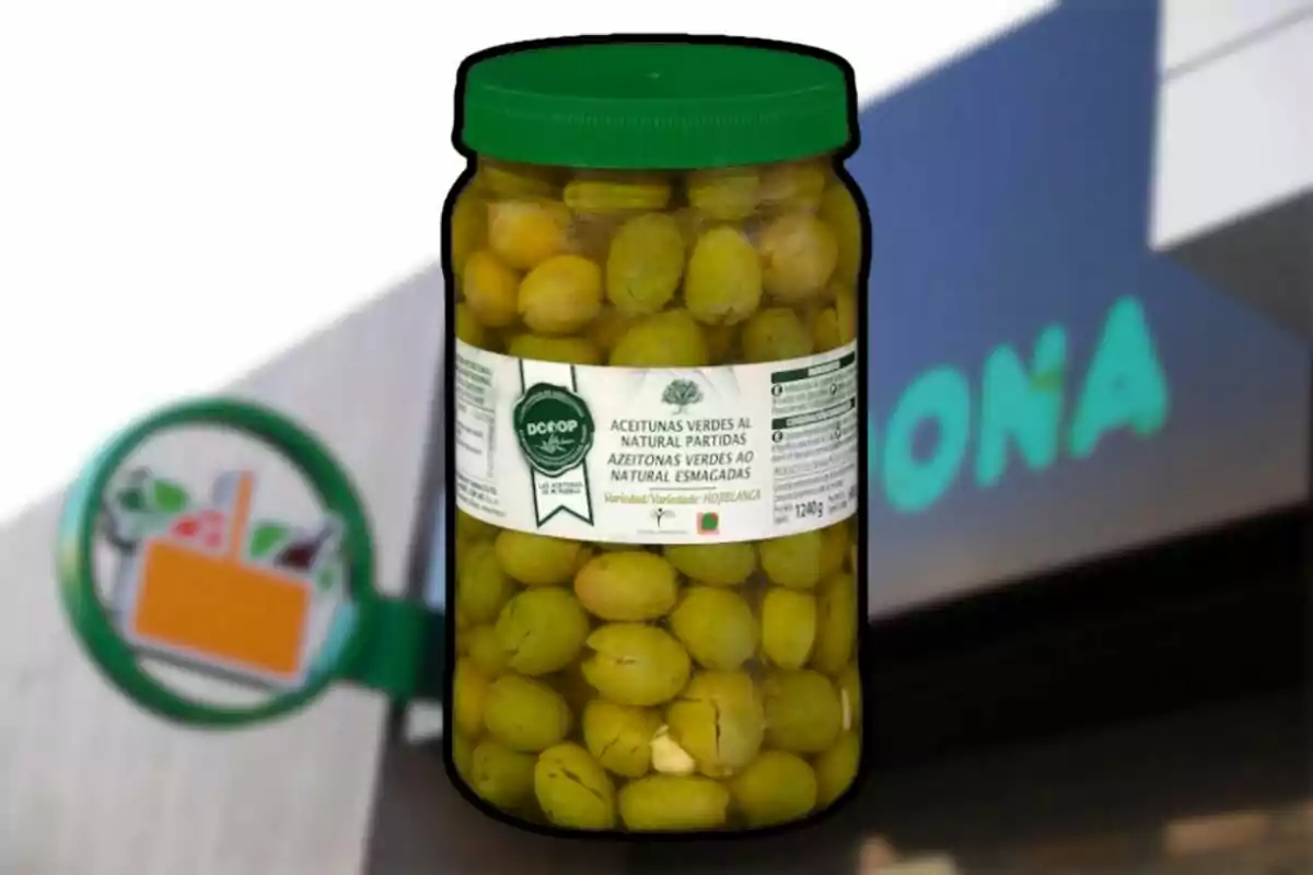 A jar of natural split green olives with a green lid and a white label against a blurred supermarket background. A jar of natural split green olives with a green lid and a white label against a blurred supermarket background.
