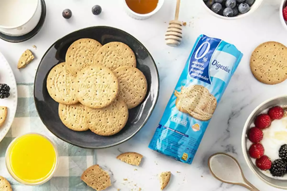 Un plato de galletas Digestive junto a un paquete de galletas, rodeado de varios alimentos como jugo de naranja, leche, miel, arándanos, frambuesas y moras. Un plato de galletas Digestive junto a un paquete de galletas, rodeado de varios alimentos como jugo de naranja, leche, miel, arándanos, frambuesas y moras.