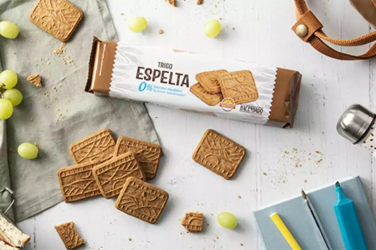 Un paquete de galletas de trigo espelta 0% azúcares añadidos está sobre una mesa blanca junto a algunas galletas sueltas, uvas verdes, una libreta con bolígrafos y una botella de agua. Un paquete de galletas de trigo espelta 0% azúcares añadidos está sobre una mesa blanca junto a algunas galletas sueltas, uvas verdes, una libreta con bolígrafos y una botella de agua.