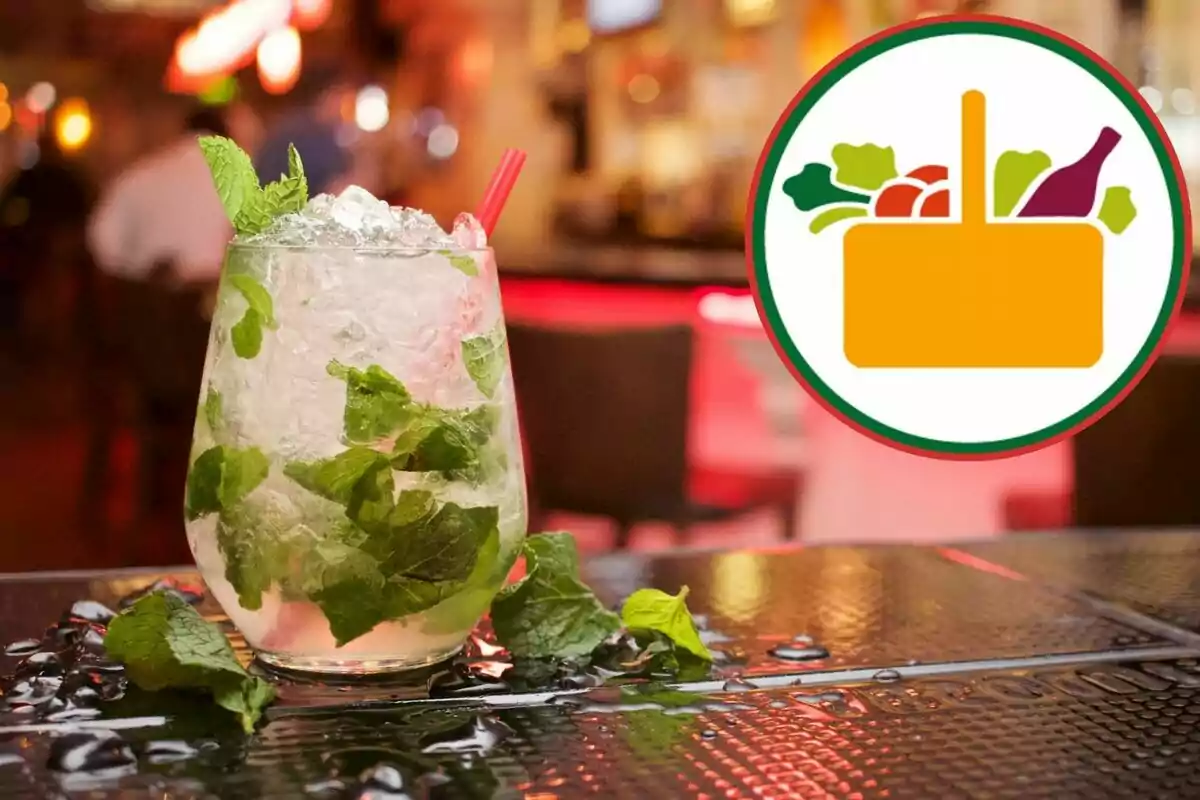 Un còctel amb fulles de menta i gel en un got sobre una taula mullada en un ambient de bar amb un logotip de cistella d'aliments al costat. Un còctel amb fulles de menta i gel en un got sobre una taula mullada en un ambient de bar amb un logotip de cistella d'aliments al costat.
