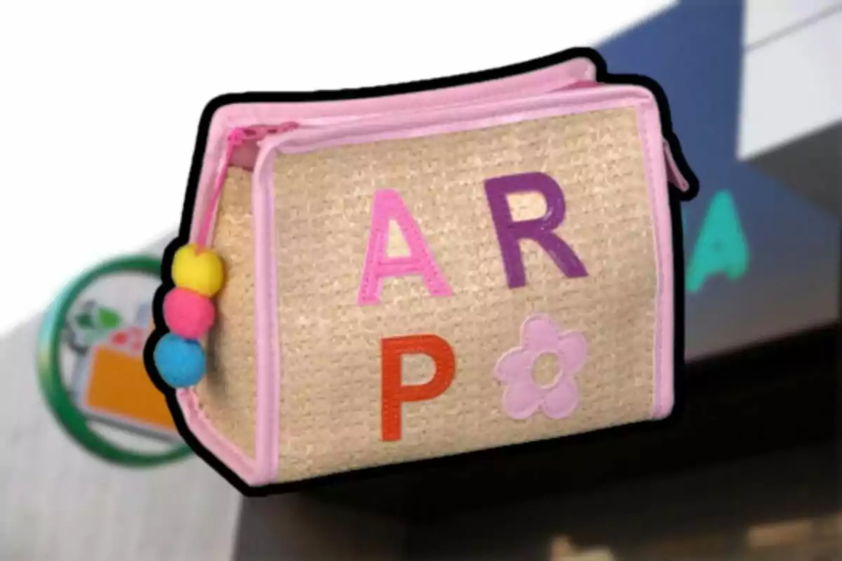Una bossa de tela beix amb vores rosades i lletres de colors, adornada amb pompons de colors en un lateral. Una bossa de tela beix amb vores rosades i lletres de colors, adornada amb pompons de colors en un lateral.