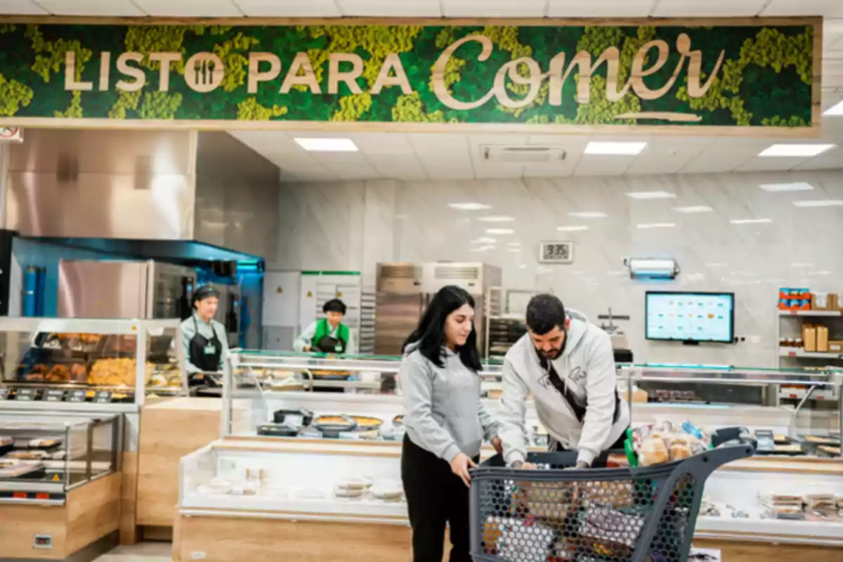 A couple with a shopping cart in a prepared food section of a supermarket with a sign that says A couple with a shopping cart in a prepared food section of a supermarket with a sign that says