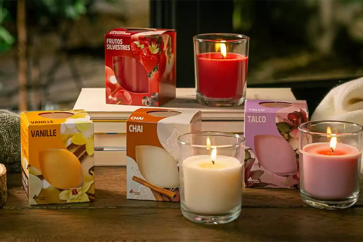 Cajas de velas aromáticas de diferentes fragancias como frutos silvestres, vainilla, chai y talco junto a velas encendidas en vasos de vidrio sobre una mesa de madera.