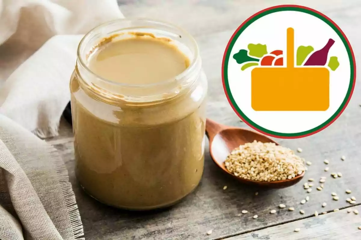 Pot de crema de sèsam sobre una taula de fusta al costat d’una cullera amb llavors i una icona de cistell d’aliments Pot de crema de sèsam sobre una taula de fusta al costat d’una cullera amb llavors i una icona de cistell d’aliments