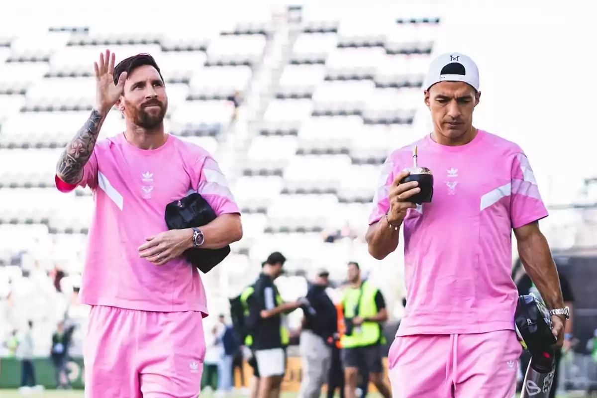 Dues persones amb roba esportiva rosa caminen en un estadi, una d'elles saluda mentre l'altra sosté un mate. Dues persones amb roba esportiva rosa caminen en un estadi, una d'elles saluda mentre l'altra sosté un mate.