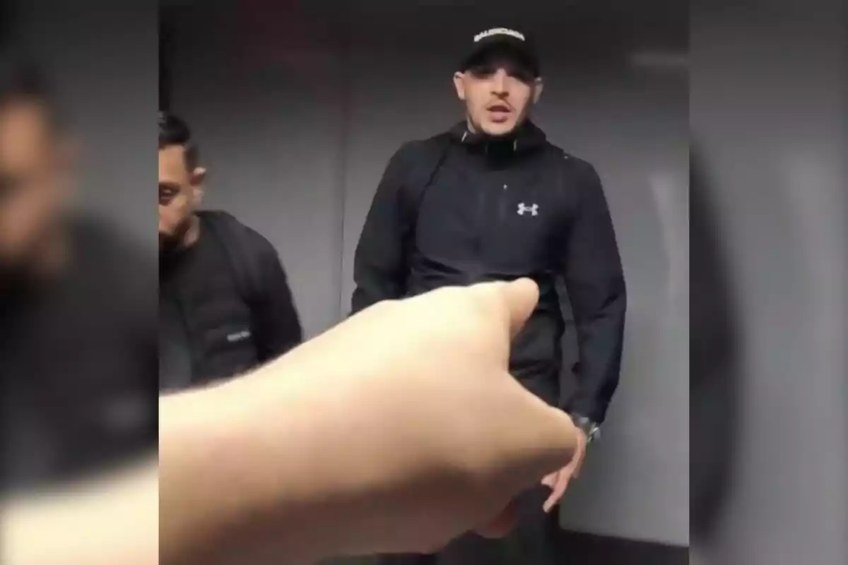 Una mà en primer pla assenyala un home amb gorra i roba esportiva negra que està dret al costat d’una altra persona en un entorn interior.