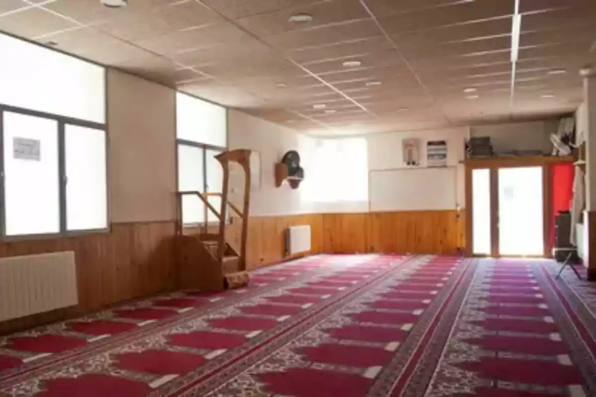 A prayer room with a red carpet and a wooden pulpit, illuminated by large windows.