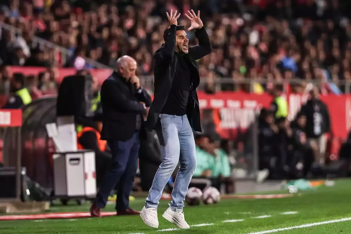 A soccer coach on the sideline of the field gestures enthusiastically as another man looks on thoughtfully in the background.