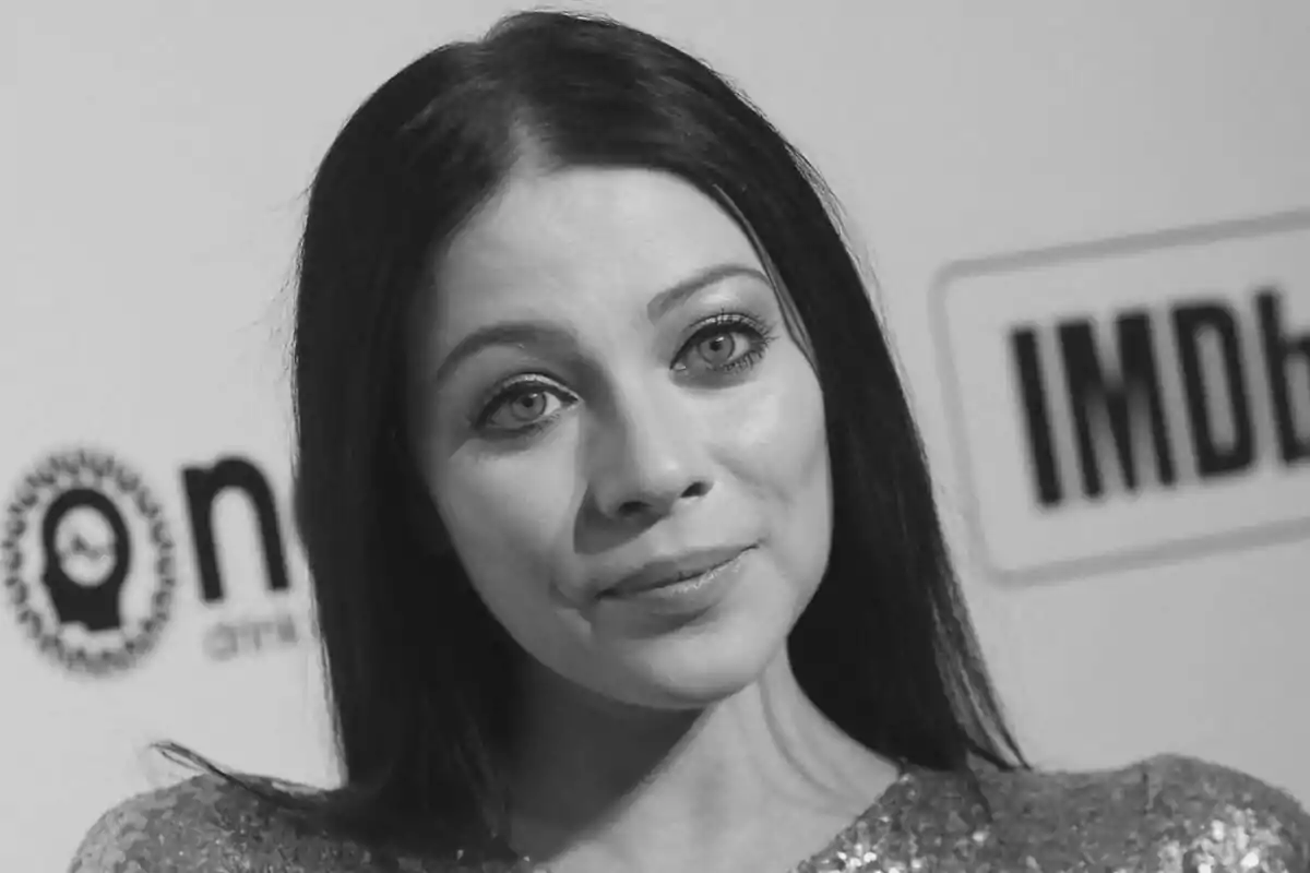 Michelle Trachtenberg amb cabell fosc i expressió neutral en un esdeveniment amb fons de logotips.