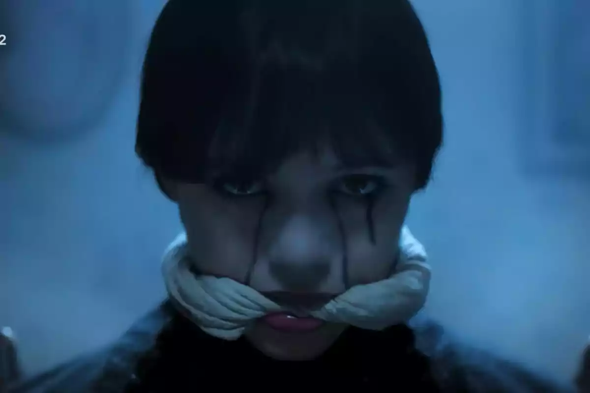 Jenna Ortega amb expressió seriosa i maquillatge fosc als ulls, amb una tela blanca lligada al voltant de la boca en un ambient de llum blavosa a la sèrie Dimecres