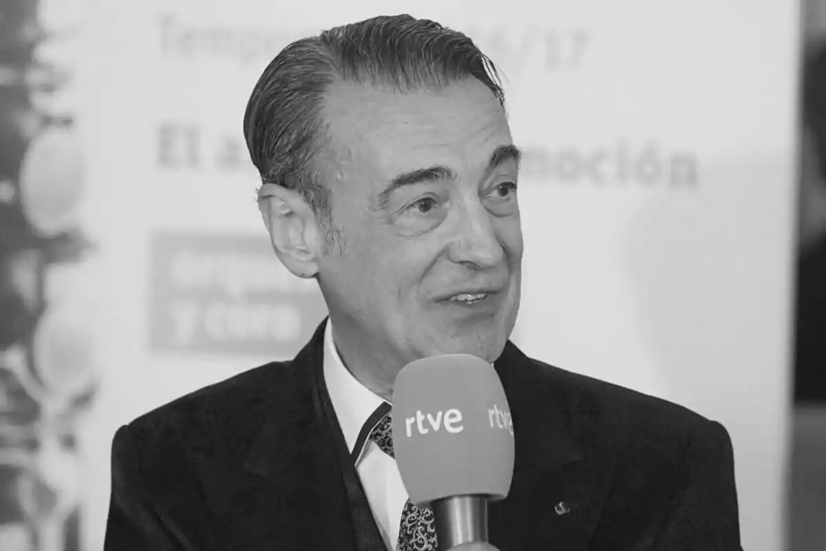 Fotografía en blanco y negro de Miguel Ángel Gómez-Martínez con traje y corbata sostiene un micrófono de RTVE mientras habla.