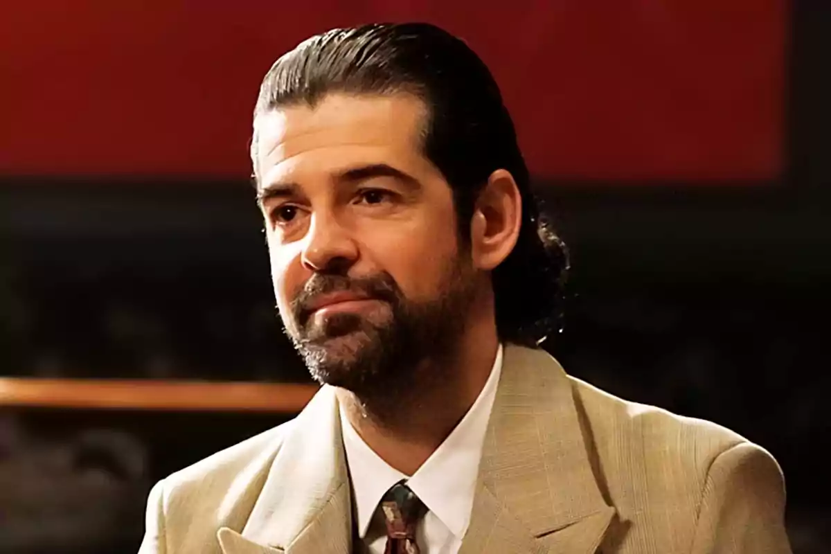 Primer pla de Miguel Ángel Muñoz amb barba i cabell fosc pentinat enrere, vestint un vestit beix i corbata, amb expressió seriosa i fons fosc en una escena de 'La Moderna'. Primer pla de Miguel Ángel Muñoz amb barba i cabell fosc pentinat enrere, vestint un vestit beix i corbata, amb expressió seriosa i fons fosc en una escena de 'La Moderna'.
