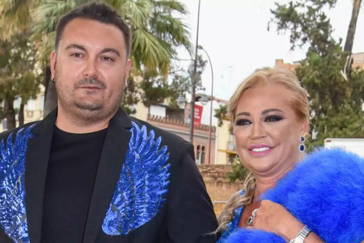 Miguel Marcos somrient en vestit negre amb ales blaves davant al costat de Belén Esteban somrient amb els cabells recollits i pendents llargs blaus
