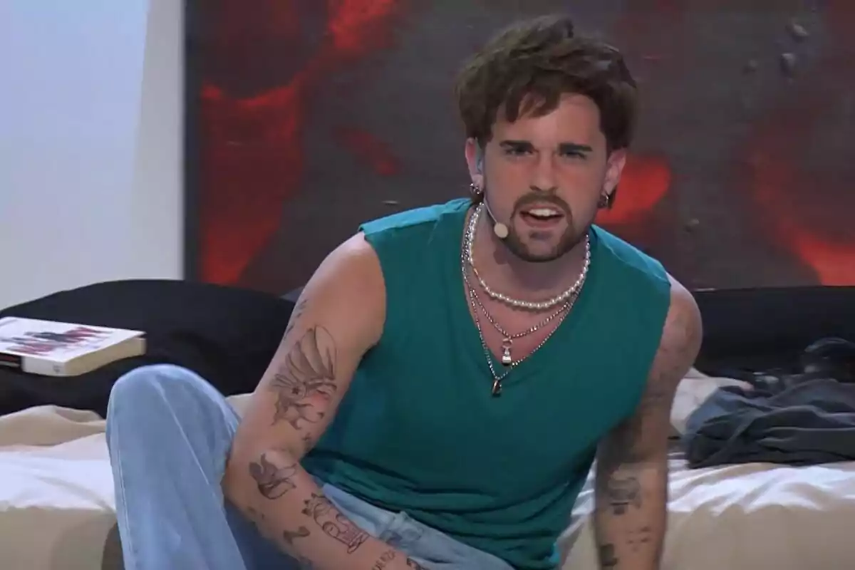 Mikel Herzog JR amb barba i tatuatges als braços està assegut en un llit, porta una samarreta de tirants verda i un micròfon de diadema a Tu Cara Me Suena. Mikel Herzog JR amb barba i tatuatges als braços està assegut en un llit, porta una samarreta de tirants verda i un micròfon de diadema a Tu Cara Me Suena.