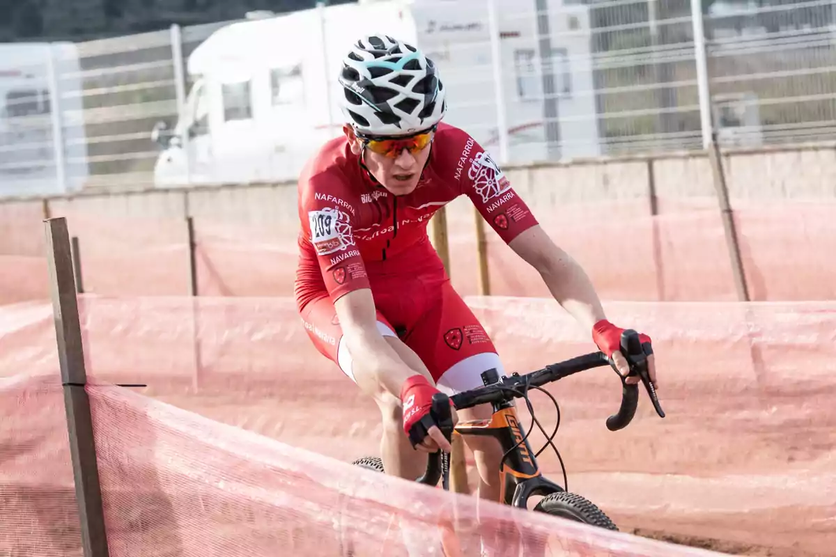 Ciclista amb uniforme vermell i casc blanc competint en una cursa de ciclisme en un circuit delimitat per xarxes roses Ciclista amb uniforme vermell i casc blanc competint en una cursa de ciclisme en un circuit delimitat per xarxes roses