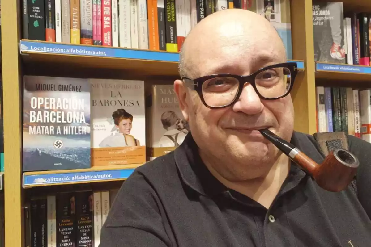 Pla curt de Miquel Giménez amb una pipa a la boca i amb una prestatgeria amb llibres de fons