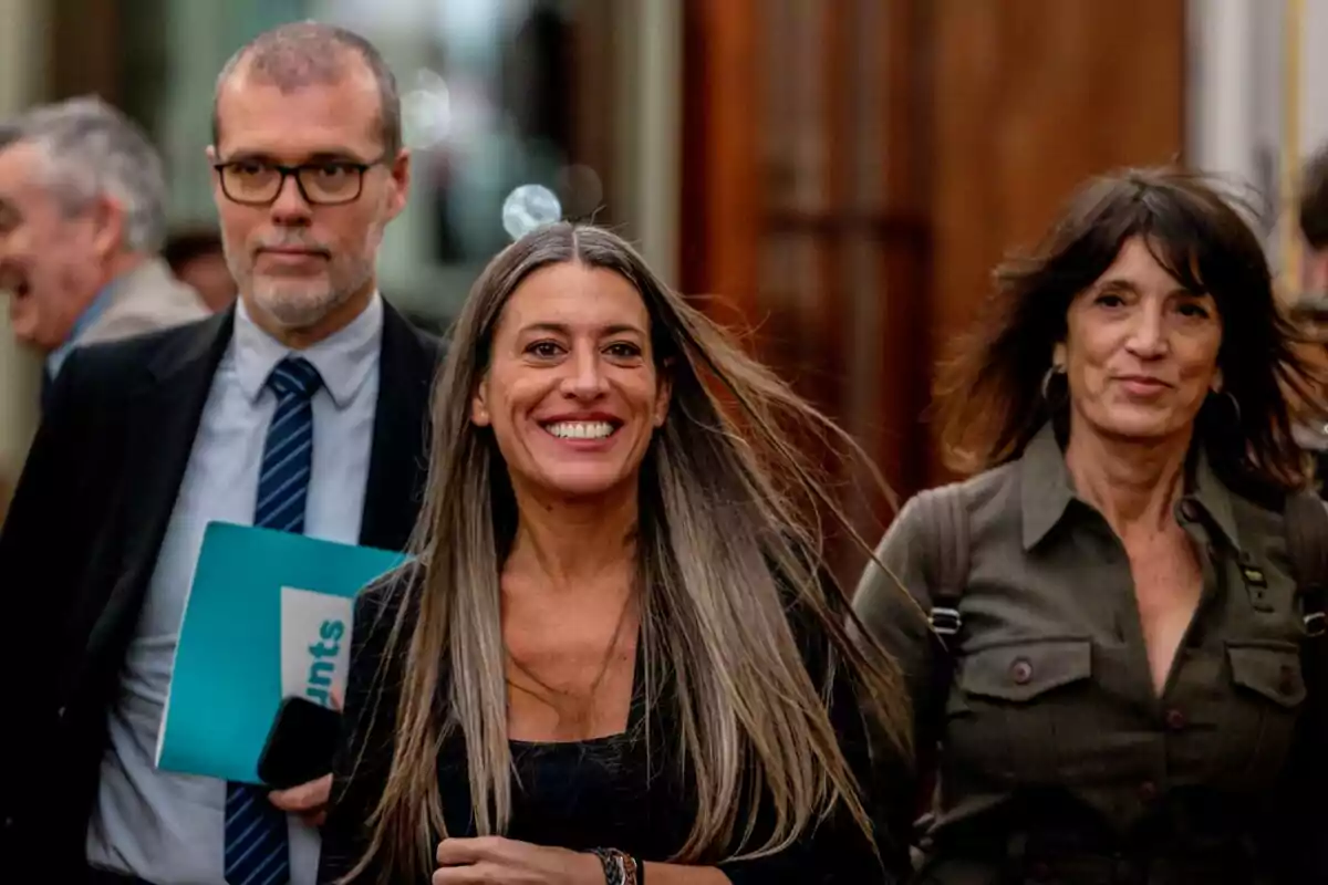 A group of people walks down a hallway, with a woman in the center standing out as she smiles while the wind moves her hair.