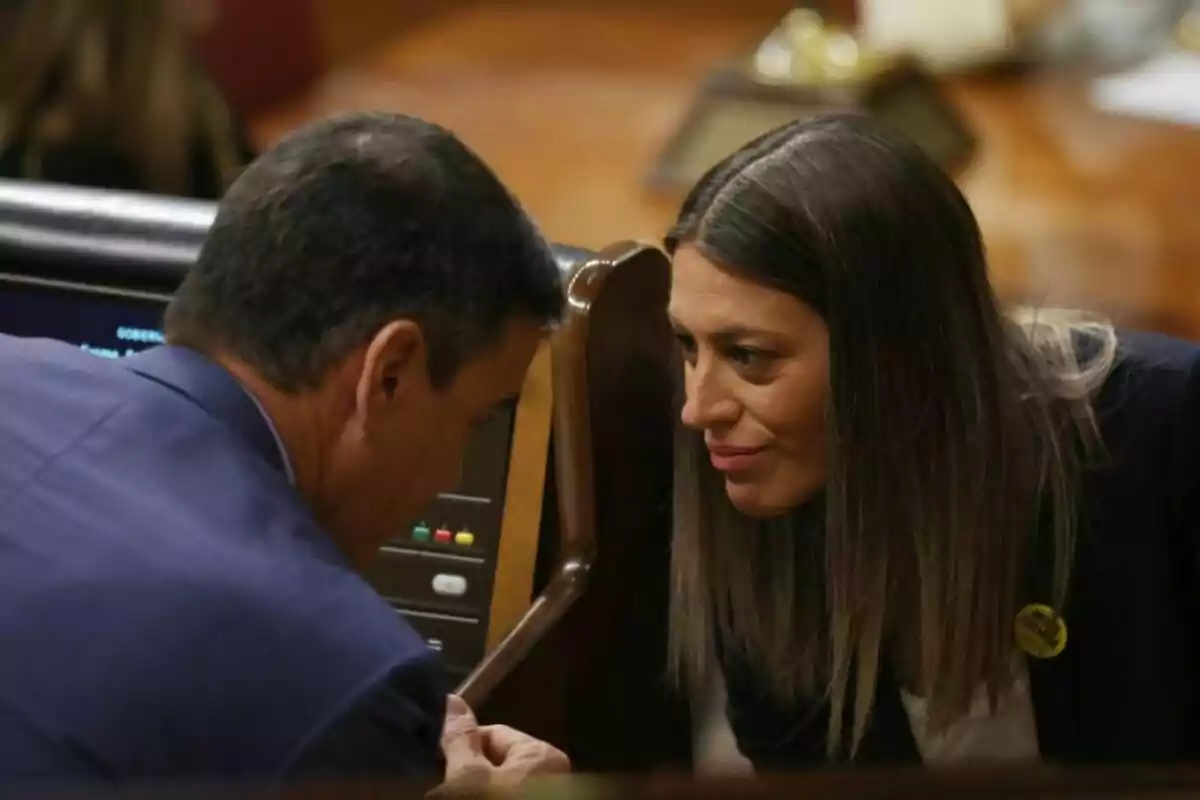 Dues persones conversen en un entorn formal, inclinades cap endavant per parlar en privat.