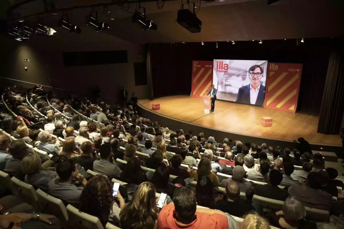 Pla general d'una sala amb desenes de persones assegudes a les grades i Salvador Illa, només davant d'un faristol a l'escenari situat a la part dreta de la imatge