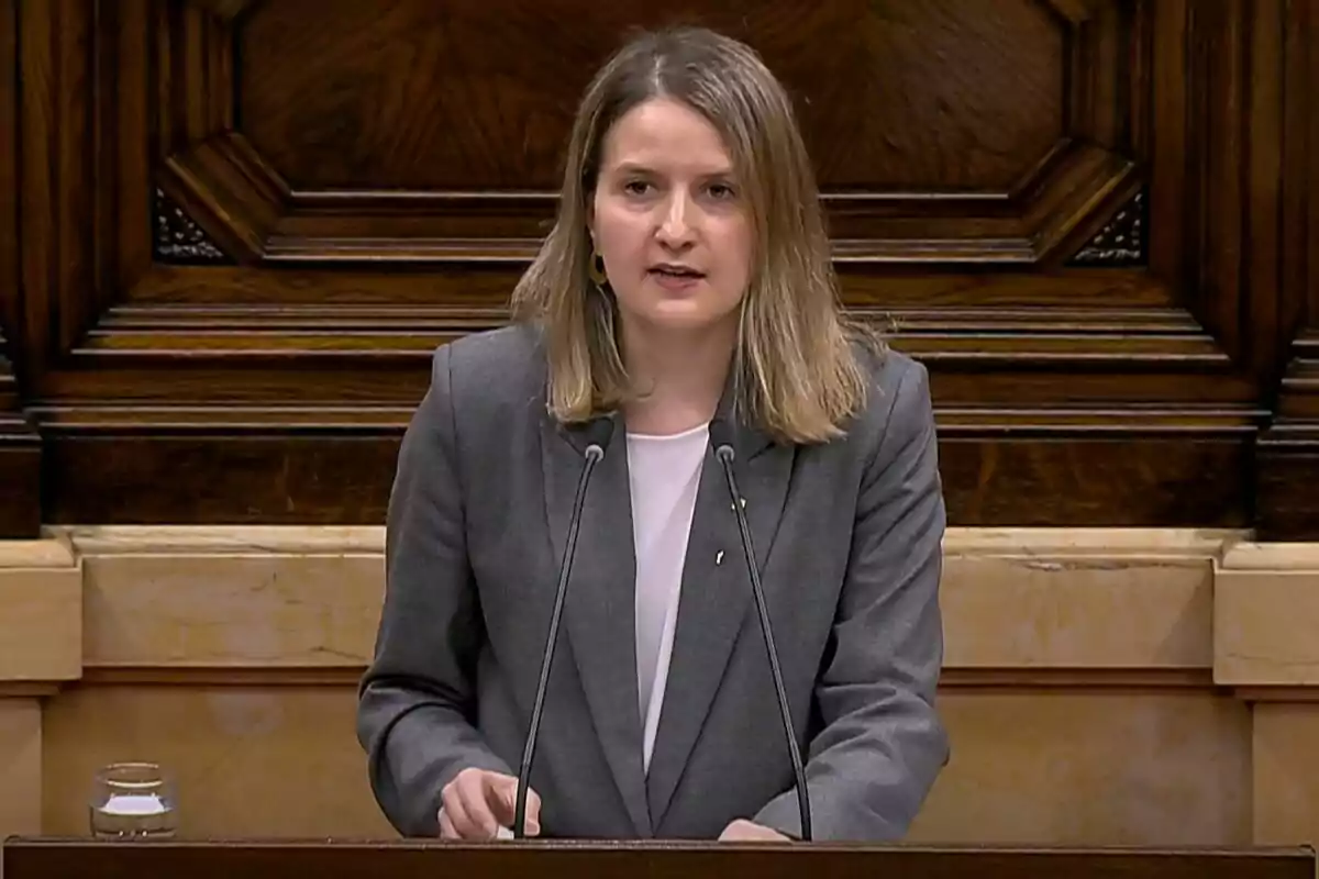 A woman speaking at a wooden podium with a microphone and a glass of water beside her.