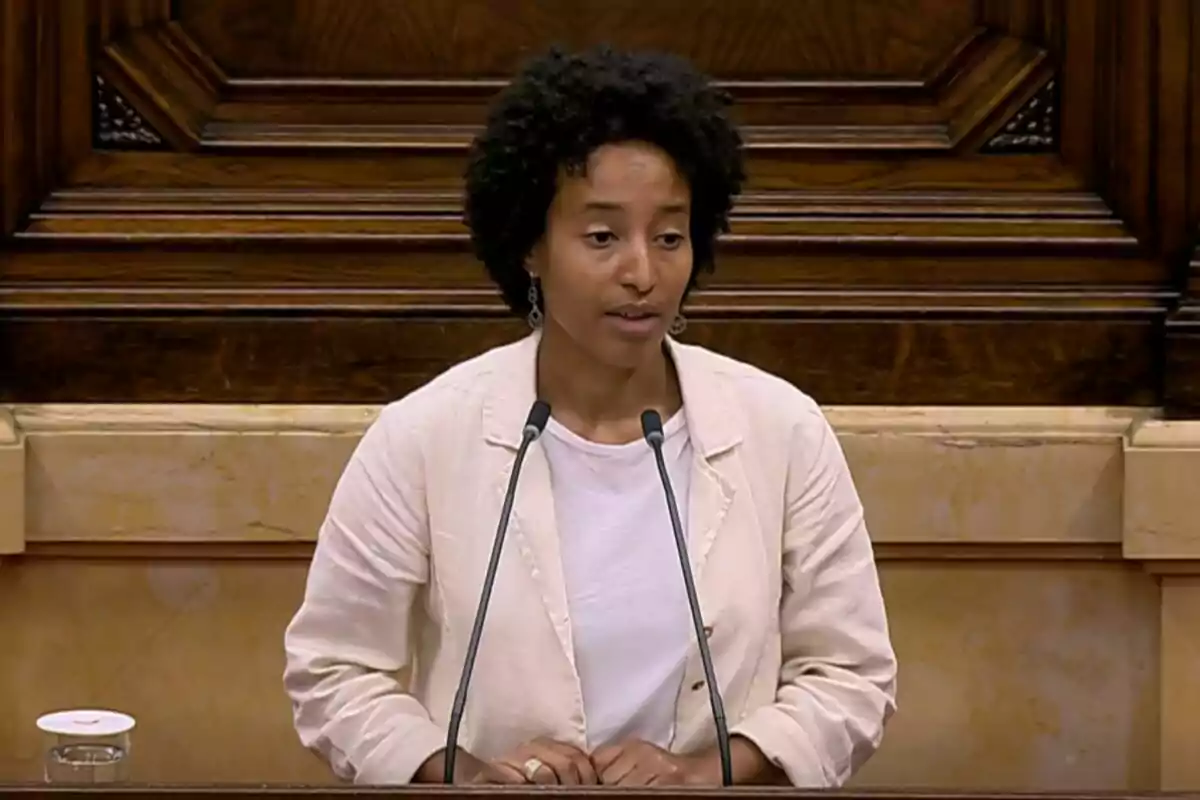 A person speaking at a podium with microphones in a wooden setting. A person speaking at a podium with microphones in a wooden setting.