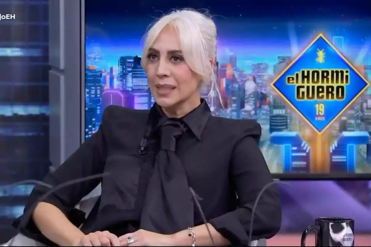 Mónica Naranjo amb cabell clar i vestida de negre està asseguda en un programa de televisió anomenat "El Hormiguero" amb un fons colorit de ciutat.