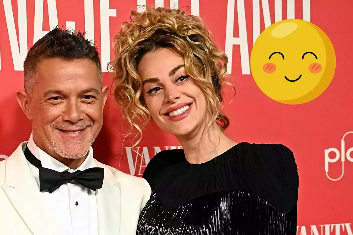 Alejandro Sanz and Candela Márquez smiling while posing in front of a red background with a smiling emoji next to them.