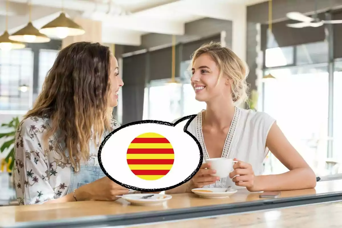 Dues dones assegudes en una cafeteria conversant i somrient, amb una bombolla de diàleg que conté una bandera amb ratlles vermelles i grogues.