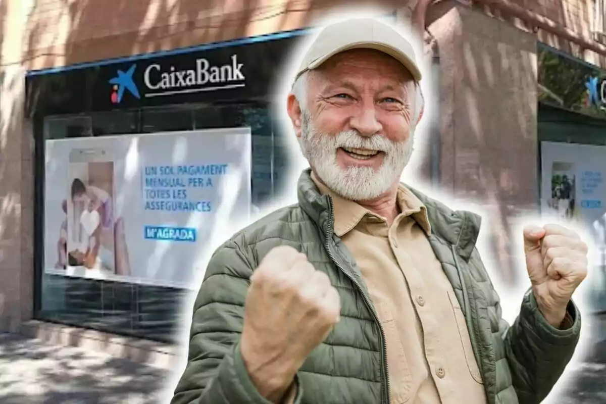 Un home gran amb barba blanca i gorra somriu i aixeca els punys en senyal de victòria davant d'una sucursal de CaixaBank.