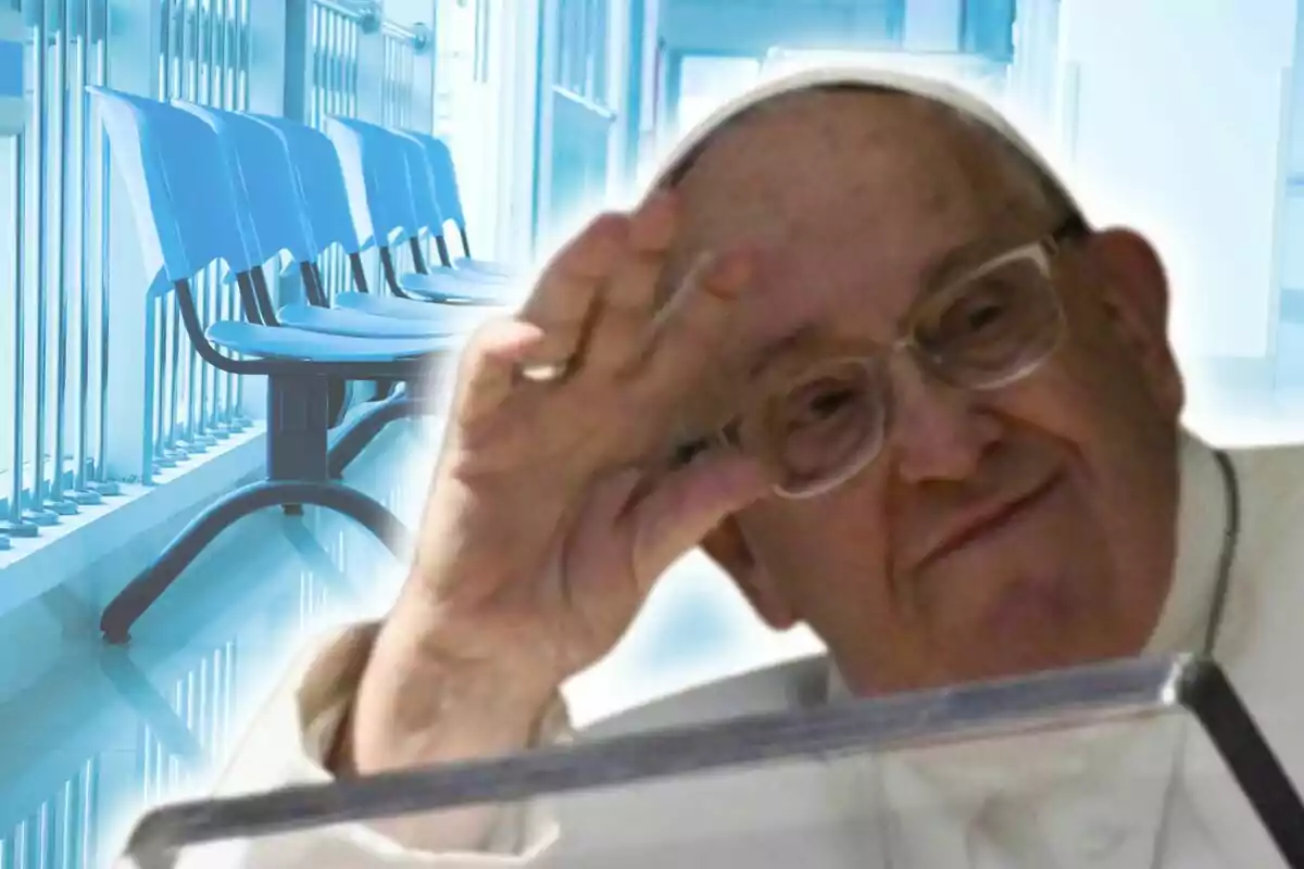 Pope Francis, wearing glasses and white attire, makes a hand gesture in front of a background of blue chairs in a hallway.