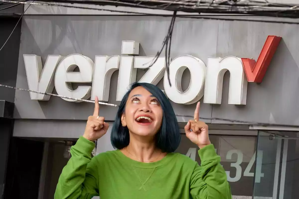 Una persona con cabello azul y suéter verde sonríe y señala hacia arriba frente a un letrero de Verizon. Una persona con cabello azul y suéter verde sonríe y señala hacia arriba frente a un letrero de Verizon.