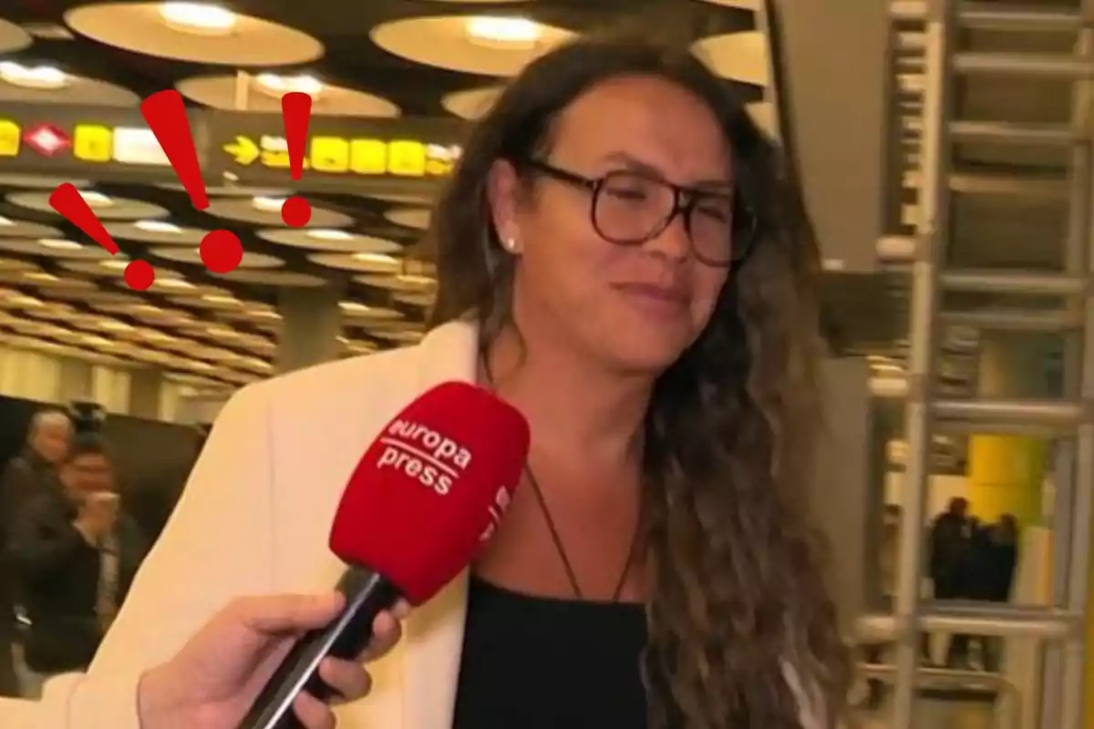 Una persona con gafas y cabello largo está siendo entrevistada en un aeropuerto, sosteniendo un micrófono de Europa Press, con signos de exclamación rojos añadidos en la imagen.