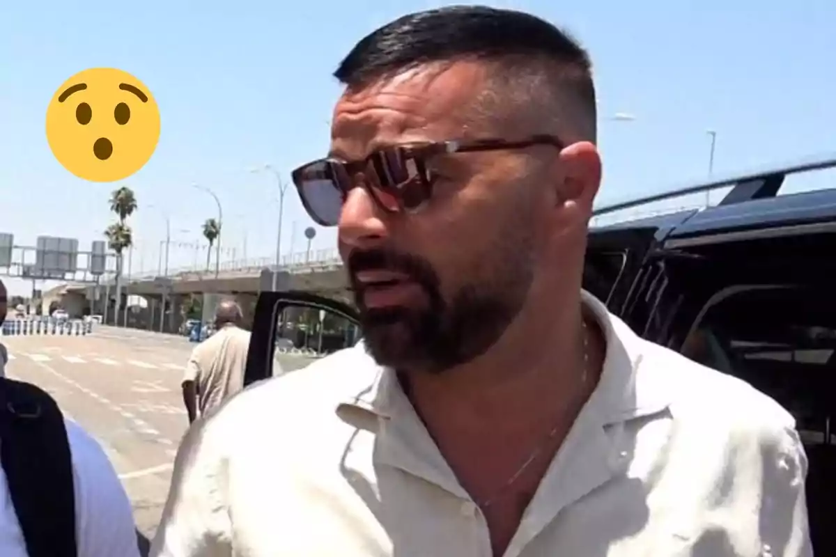 Ricky Martin with sunglasses and a beard is standing next to a vehicle in an urban setting with a clear sky.