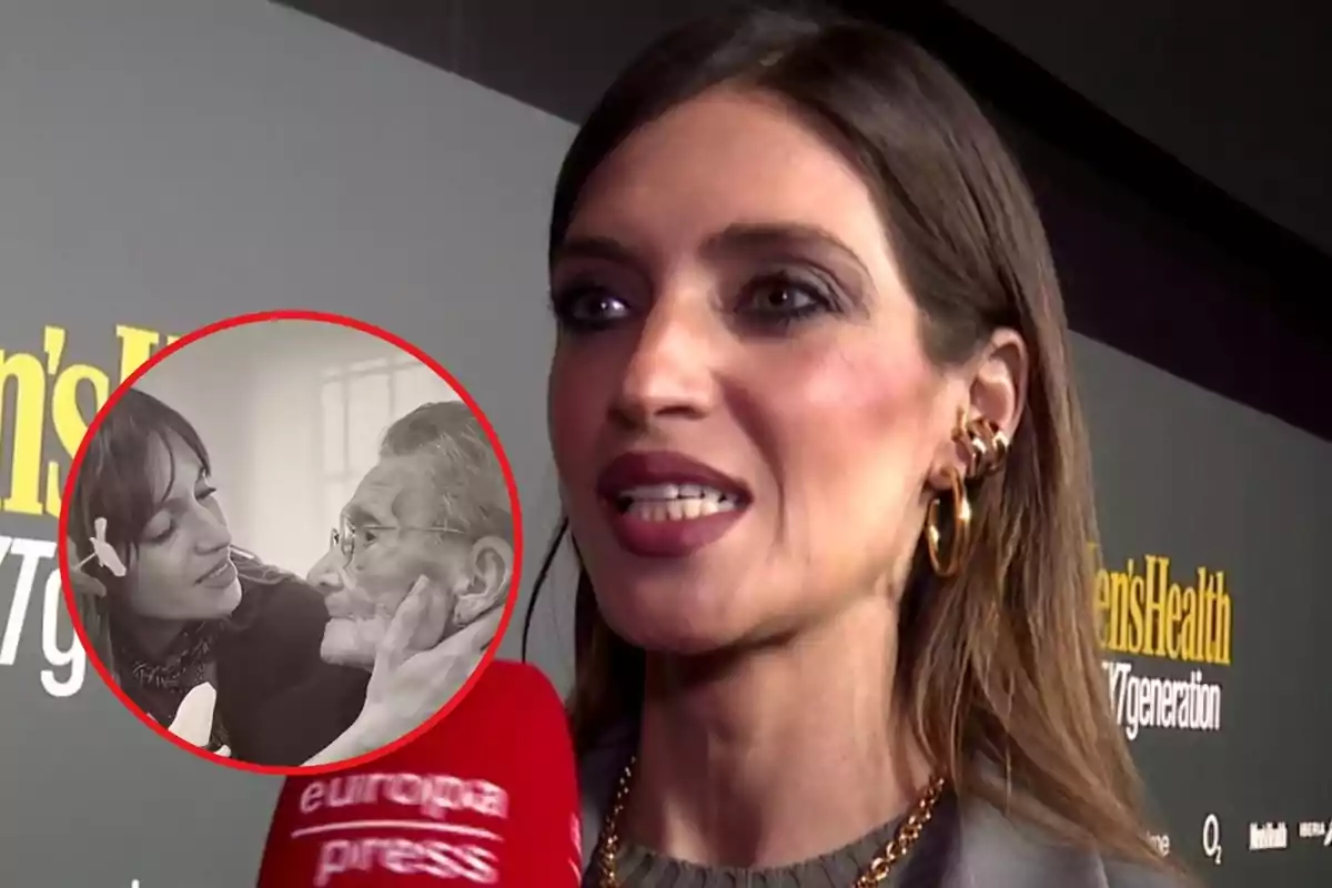 A woman with brown hair and gold earrings speaks in front of a red Europa Press microphone, with a background showing a Men's Health poster; in a circular inset, there is a black and white image of a young woman caressing the face of an older person.