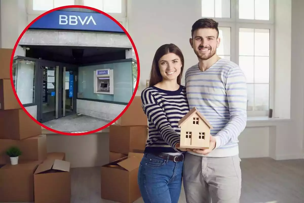 Una parella somrient sosté una casa en miniatura en una habitació amb caixes de mudança, amb una imatge inserida d?una sucursal del banc BBVA.