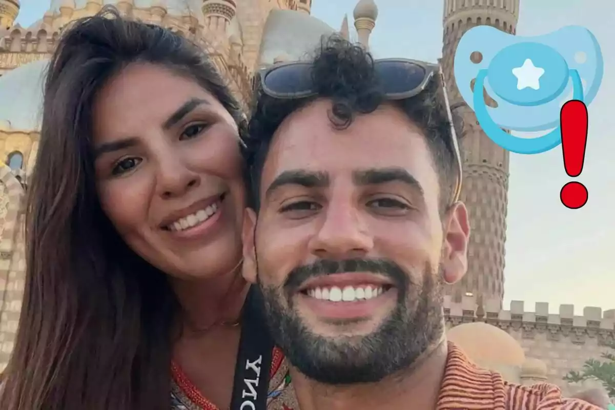 Isa Pantoja and Asraf Beno, smiling, take a selfie in front of a building with detailed architecture, while a drawing of a blue pacifier with a red exclamation mark appears in the upper right corner.