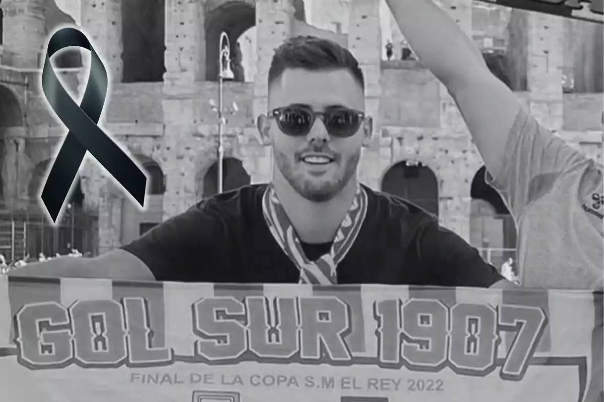 Un home amb ulleres de sol sosté una bufanda que diu "Gol Sud 1907" davant d'un edifici antic, amb un llaç negre superposat a la imatge.