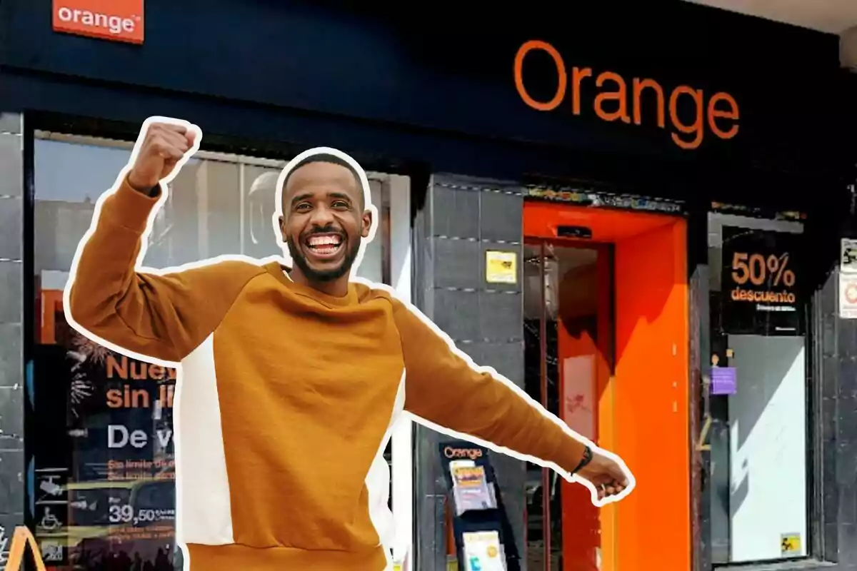 Un hombre sonriente con un suéter marrón levanta el puño frente a una tienda de Orange.