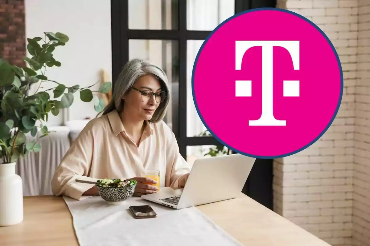 Woman with gray hair and glasses working on a laptop while holding a glass of juice, with a bowl of salad and a mobile phone on the table, next to a large pink logo. Woman with gray hair and glasses working on a laptop while holding a glass of juice, with a bowl of salad and a mobile phone on the table, next to a large pink logo.