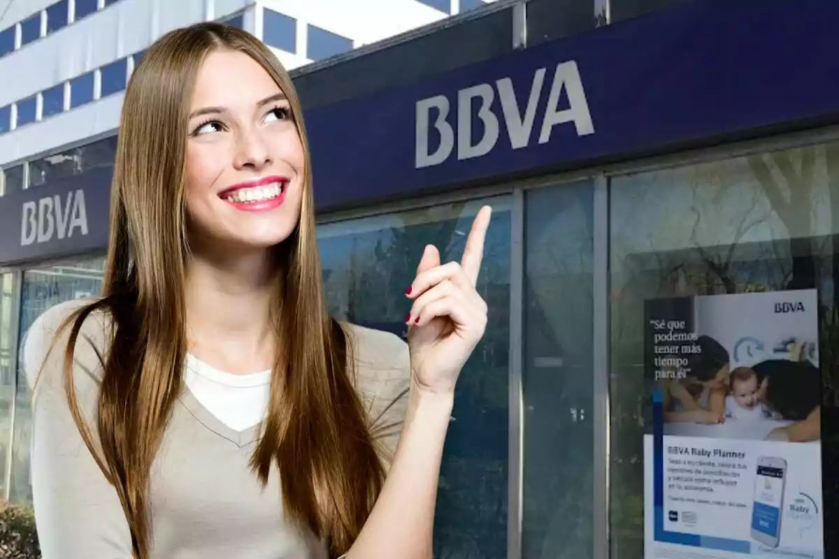 Una dona somrient amb cabells llargs i lacio assenyala amb el dit índex cap al rètol d'una sucursal de BBVA.