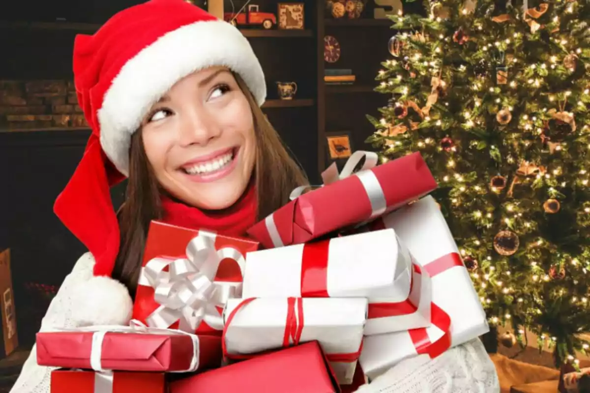 Persona sonriente con gorro de Santa sosteniendo regalos frente a un árbol de Navidad iluminado.