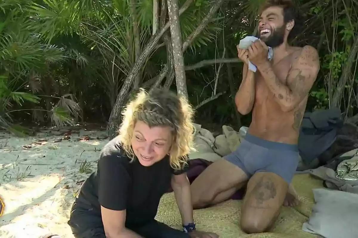 Dues persones en una platja de Supervivientes, Terelu Campos asseguda a la sorra i Montoya dreta rient, envoltades de vegetació.